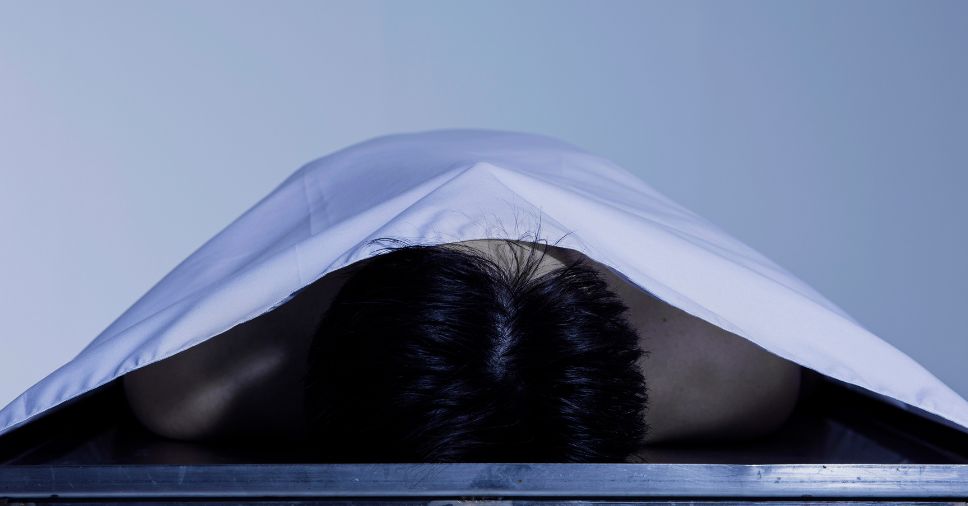 Corpo coberto por lençol branco em necrotério ilustra a decomposição e putrefação.